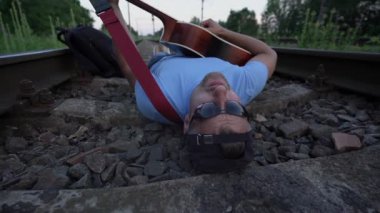 Güneş gözlüklü ve şapkalı erkek demiryollarında yatıyor gitar çalıyor ve şarkı söylüyor.
