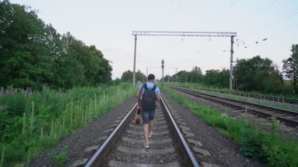 男性の旅行者はレールを歩いてバックパックとギターのリアビューを運び — ストック動画