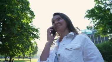 Beyazlı kadın ayakta duruyor ve parkta akıllı telefonla konuşuyor.