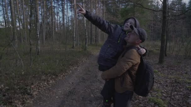 Ojciec trzyma syna w ramionach i opowiada mu o naturalnych zjawiskach w drewnie. — Wideo stockowe