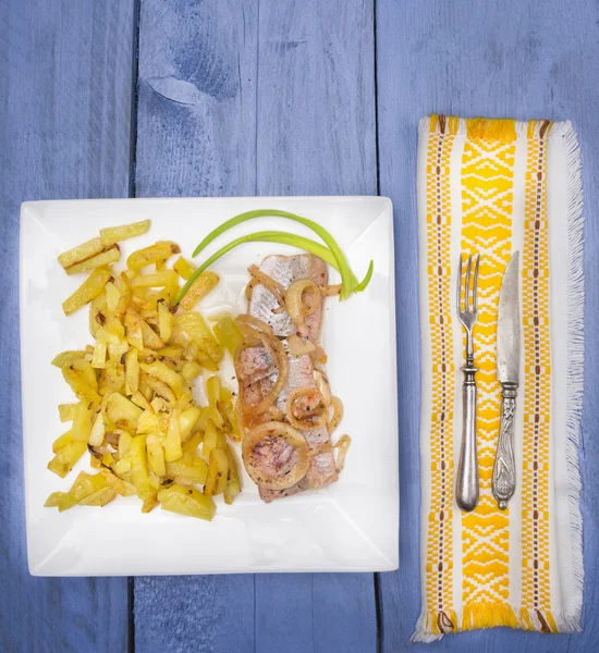 油煎的土豆鲱鱼和洋葱在白色方形板、 黄色的布、 刀和叉，服务蓝色木制的桌子上 — 图库照片