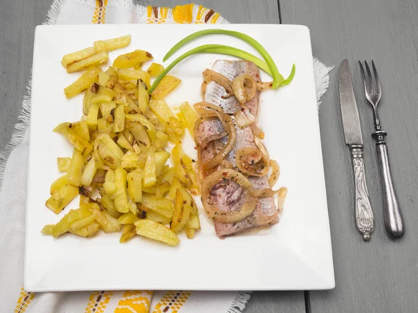 油煎的土豆鲱鱼和洋葱在老刀与叉在灰色的木桌上的白色方形板 — 图库照片