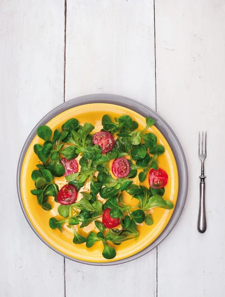 Ensalada de Feld verde dieta con tomates, sirviendo en plato pesado amarillo y gris sobre mesa de madera blanca —  Fotos de Stock