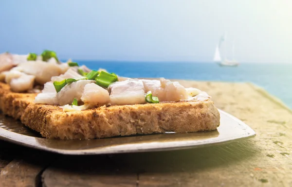 Toast with salted herring and onion in metal tray, on wooden table, against blue sea and blue sky — Stock Photo, Image