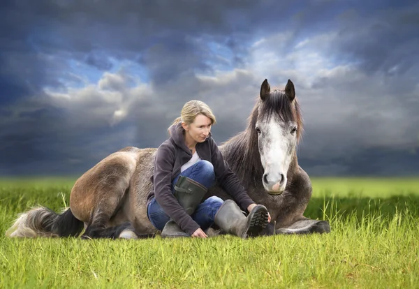 Кінь і жінка лежать в зеленому полі проти неба після шторму, милосердя, походження — стокове фото