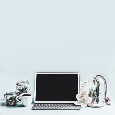 Dizüstü bilgisayar ya da defter, açık mavi masaüstünde bir fincan kahve ve çiçekli sulu bitkilerle makara yapıyor. Ev işleri bürosu çalışma alanı. Tasarımınız için siyah boş ekran