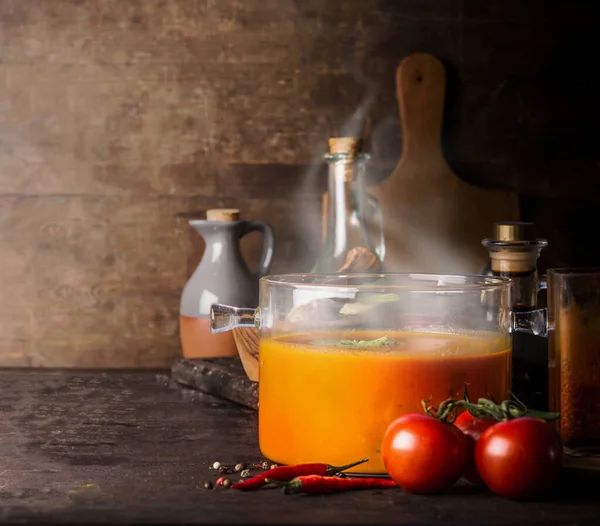 Glass cooking pot with hot tomato soup and blow off steam standing on rustic kitchen table with ingredients. Copy space. Healthy eating and homemade cooking