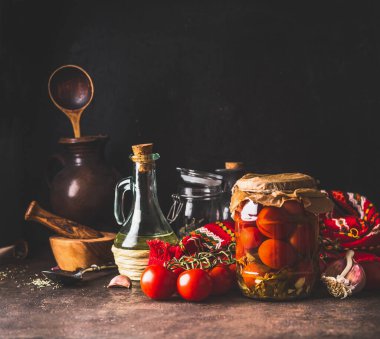 Home canned pickled tomatoes in glass jar on rustic table with ingredients. Homemade harvest storage. Copy space. Healthy food.