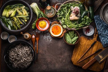 Asian food background. Tasty vegetarian ingredients. Various green vegetables,  soba noodles - buckwheat noodles, flavor salt and chili hot sauce on rustic kitchen table background with chopsticks