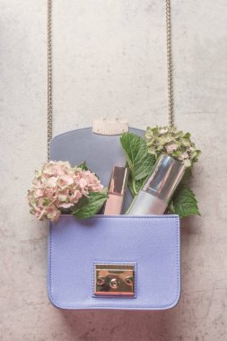 Açık gri beton masa üzerinde ortanca çiçekleri ve kozmetik ürünlerle dolu mor bir çanta. Yaşam tarzı konsepti. Üst görünüm.