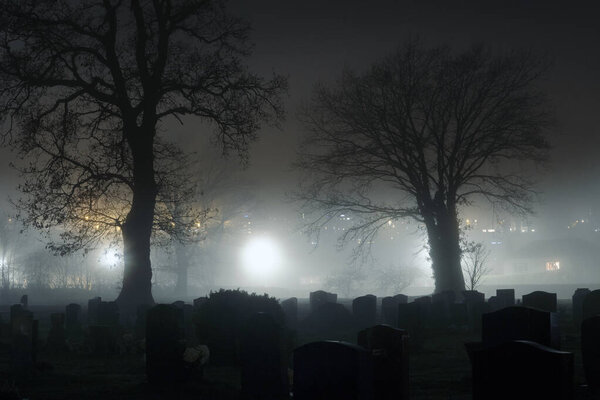 A spooky concept of a cemetery on a foggy winters night. With graves silhouetted by street lights. 