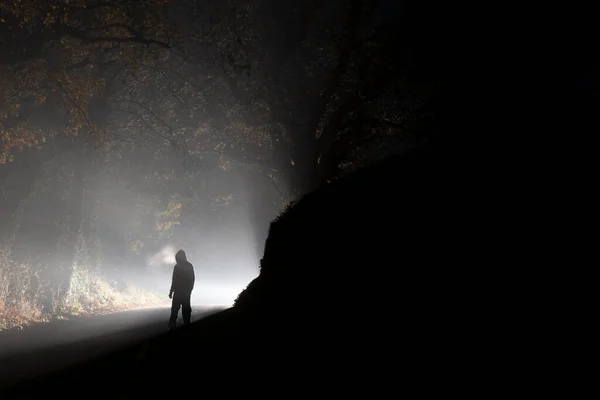 A mysterious hooded figure silhouetted by car headlights standing on a road on a spooky foggy winters night.