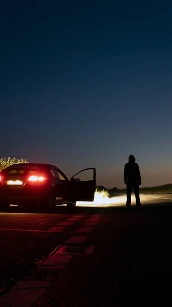 一个神秘的头戴头巾的人晚上站在一辆停放的汽车旁边 在一条可怕的空旷乡间道路上 具有垂直格式 — 图库照片