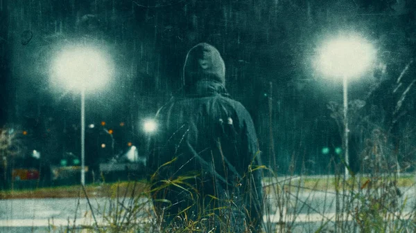 A dark grunge edit of a mysterious hooded figure, back to camera. On spooky street. On a winters night with rain.
