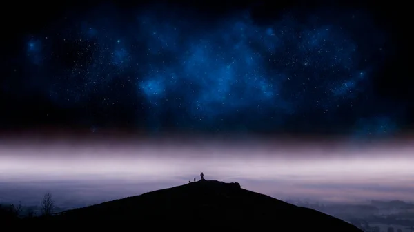 A mystical concept of a man standing on a hill with dogs. Silhouetted against a night sky of a universe of stars. With a misty landscape below.