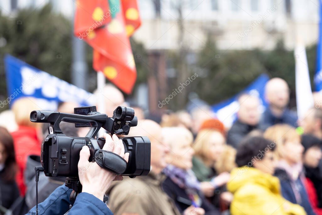 Filming street protest, television camera lens in the focus, blurred people in the background