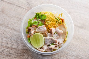 Dry noodles pork with red roasted pork and vegetable in plastic bowl on wooden table, top view Thai and China Asian food yellow noodles lemon lime