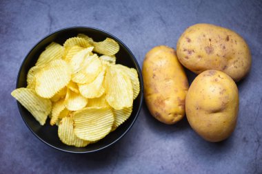 Mutfak masasında çıtır patates cipsi, arka planda taze çiğ patates, kasede patates cipsi.