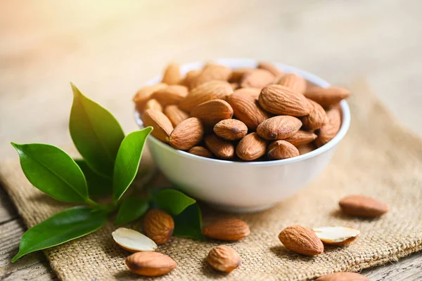 Almonds nuts on white bowl and green leaf on sack background, Delicious sweet almonds on the wooden table , roasted almond nut for healthy food and snack