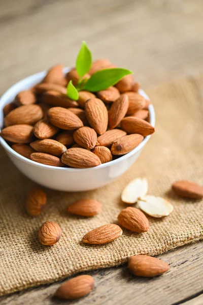 Delicious sweet almonds on the wooden table, roasted almond nut for healthy food and snack, Almonds nuts on white bowl and green leaf on sack background