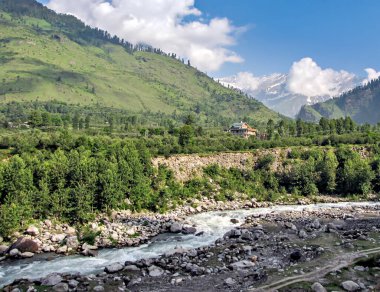 Beas nehrinin kıyısında güzel, yalnız bir ev. Karla kaplı dağ, mavi bulutlar ve arka planda yeşillik.