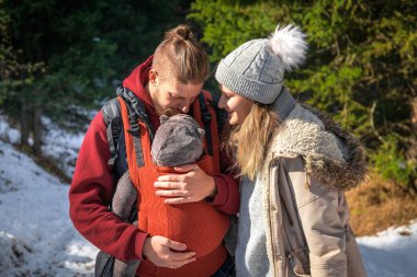 Young caucasian babywearing family with their baby in baby carrier winter outdoor adventure. clipart