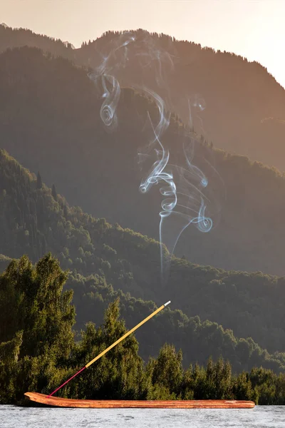 Burning stick of incense stands on a wooden stand on the background of the sunset in the mountains. Vertical, copy space