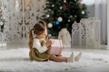 Little girl holding new year gift while sitting on the carpet in christmas interior