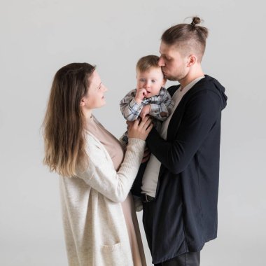 Bir adam çocuğunu kollarına alır ve onu öperken karısı beyaz bir arka planda onlara bakar..