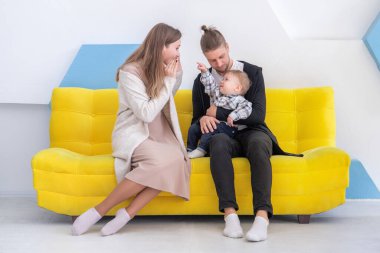 Sarışın ve çocuklu genç bir aile, parlak bir odadaki sarı bir kanepede eğleniyor..
