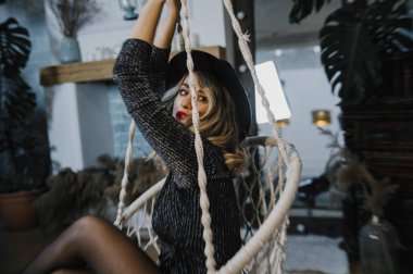 Kısa siyah elbisesi, şapkası ve ayakkabıları olan güzel, seksi, duygusal bir kız bir partide şık bir stüdyoda poz veriyor.