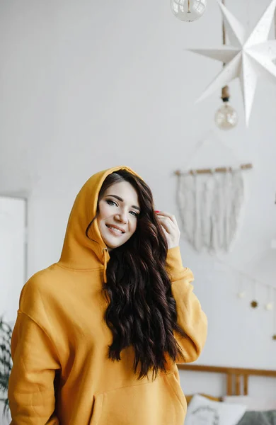 可爱的黑发姑娘 穿着黄色的帽衫 摆着吹风机的姿势 笑着看美容院的广告 — 图库照片