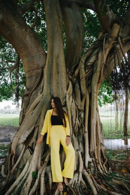 Sarı takım elbiseli ince esmer bir kız Dominik Cumhuriyeti 'ndeki büyük bir ağacın yanındaki tropikal bir adada zarif bir şekilde golf kulübüne poz veriyor.