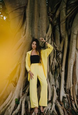 Sarı takım elbiseli ince esmer bir kız Dominik Cumhuriyeti 'ndeki büyük bir ağacın yanındaki tropikal bir adada zarif bir şekilde golf kulübüne poz veriyor.