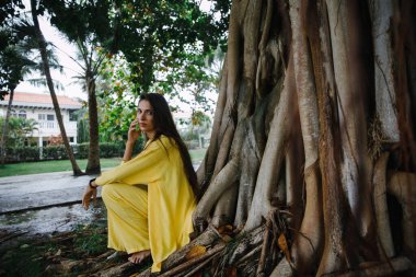 Sarı takım elbiseli ince esmer bir kız Dominik Cumhuriyeti 'ndeki büyük bir ağacın yanındaki tropikal bir adada zarif bir şekilde golf kulübüne poz veriyor.