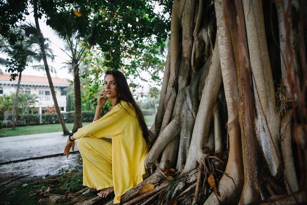 Sarı takım elbiseli ince esmer bir kız Dominik Cumhuriyeti 'ndeki büyük bir ağacın yanındaki tropikal bir adada zarif bir şekilde golf kulübüne poz veriyor.
