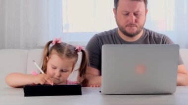 Bir iş adamının üzgün babası evinde, tablette oynayan 3 yaşındaki küçük bir kızın yanında dizüstü bilgisayarla çalışıyor. Modern aile. Serbest meslek kavramı. Aygıtlara bağımlılık