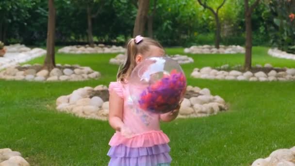 Обиженная грустная маленькая девочка, 3 года, с двумя хвостиками на голове, одетая в тонкое и разноцветное платье розово-голубого цвета, играет с ярким прозрачным мячом с разноцветной — стоковое видео
