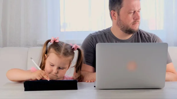Ledsen far till en affärsman arbetar på en bärbar dator hemma bredvid en liten flicka, 3 år gammal, spelar i tabletten. Modern familj. Frilansbegreppet. Beroende av prylar — Stockfoto