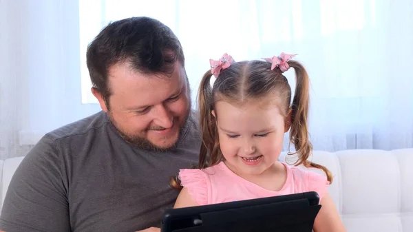 3 살짜리 여자 아이를 웃으며 아버지와 함께 타블렛으로 공부하고 집에서 온라인으로 강의를 보고 있습니다. 아버지는 딸의 학업을 도와 줍니다. 온라인 학교의 거리 학습 개념. — 스톡 사진