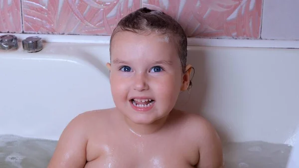 Petite fille rieuse de 3 ans se baigne et se lave avec de la mousse sur la tête dans l'eau dans la salle de bain à la maison. Bébé concept de soins du corps, hygiène. — Photo