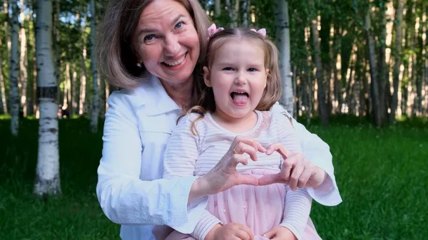 Laughing grandmother and granddaughter, 3 years old, show the heart out of their hands. The concept of a happy relationship between generations, raising children — Stock Photo, Image