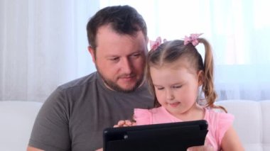 Üç yaşında komik küçük bir kız ve babası birlikte tablet okuyor ve evde online bir ders izliyorlar. Babam kızına çalışmasında yardım ediyor. Çevrimiçi okul uzaklığı öğrenme kavramı.