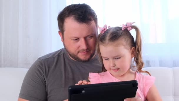 Ein lustiges kleines Mädchen im Alter von 3 Jahren und ihr Vater lernen zusammen auf einem Tablet und schauen sich zu Hause eine Unterrichtsstunde online an. Papa hilft seiner Tochter beim Studium. Online-Fernstudienkonzept für Schulen. — Stockvideo