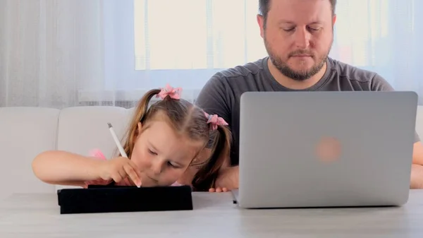 Pai triste de um homem de negócios está trabalhando em um laptop em casa ao lado de uma menina, 3 anos, jogando no tablet. Família moderna. Conceito freelance. Vício em gadgets — Fotografia de Stock