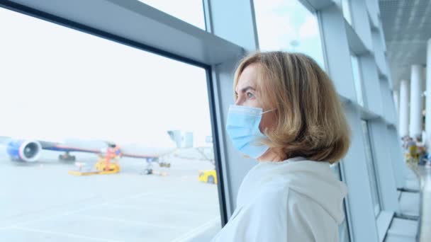 Adulto Donna con maschera protettiva è in piedi alla finestra nel terminal dell'aeroporto in attesa della partenza di un volo a causa di restrizioni di viaggio a causa della pandemia coronavirus, un anziano di 50 anni — Video Stock