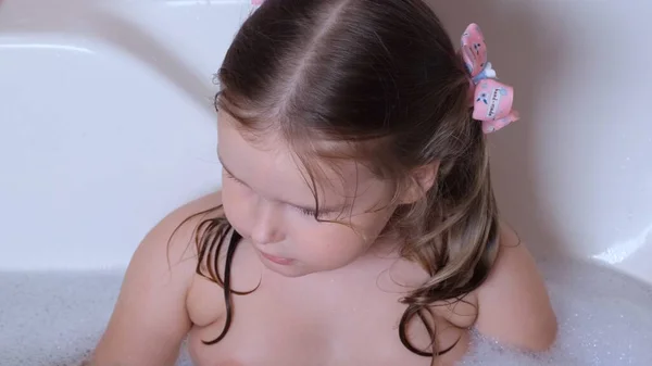 Pequena menina bonito 3 anos de idade, com dois rabos de cavalo na cabeça, banhos e lavagens na água no banheiro em casa. Conceito de cuidados com o corpo do bebê, higiene. — Fotografia de Stock