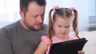 Üç yaşında komik küçük bir kız ve babası birlikte tablet okuyor ve evde online bir ders izliyorlar. Babam kızına çalışmasında yardım ediyor. Çevrimiçi okul uzaklığı öğrenme kavramı.