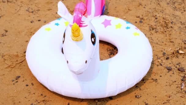 Uppblåsbar ring i form av en vit enhörning på sanden på stranden. Fantasy simring för sommaren segling till sjöss. Dröm koncept för sommarsemester, solig dag och varmt väder. — Stockvideo