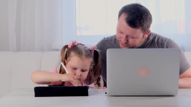 Милый отец бизнесмена работает на ноутбуке у себя дома рядом с маленькой девочкой, 3 года, играет в планшете. Современная семья. Концепция фрилансера. Зависимость от гаджетов — стоковое видео